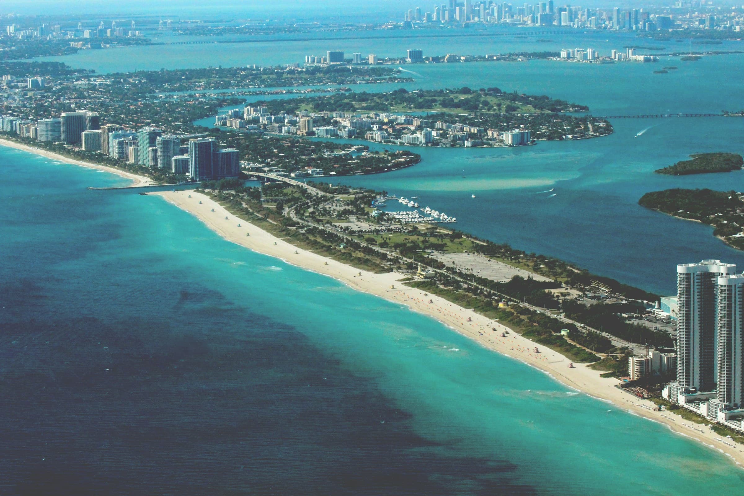Bright daytime aerial of Miami Beach, turquoise Atlantic water, and the downtown Miami skyline.